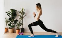 Side view of fit lady in sports clothes and wristwatch standing in Anjaneyasana pose while practicing yoga on mat near dumbbells and potted plants at home and looking away